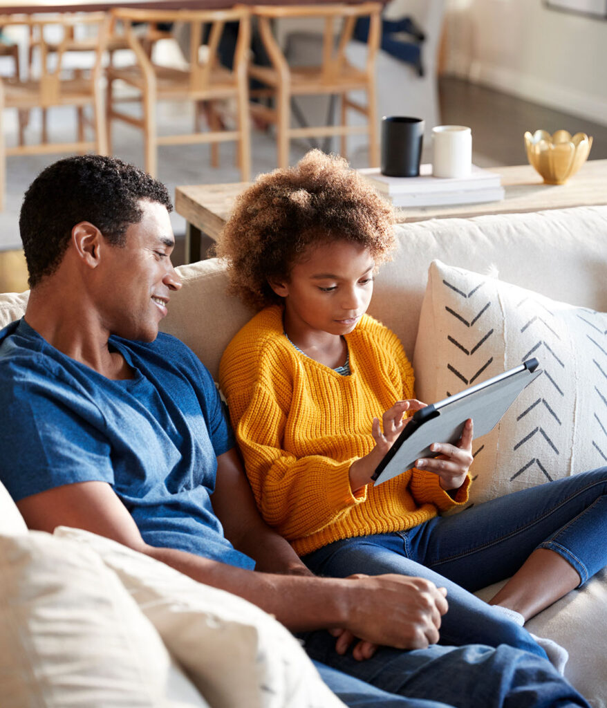 A man sitting on a couch with his daughter as she plays on a tablet.